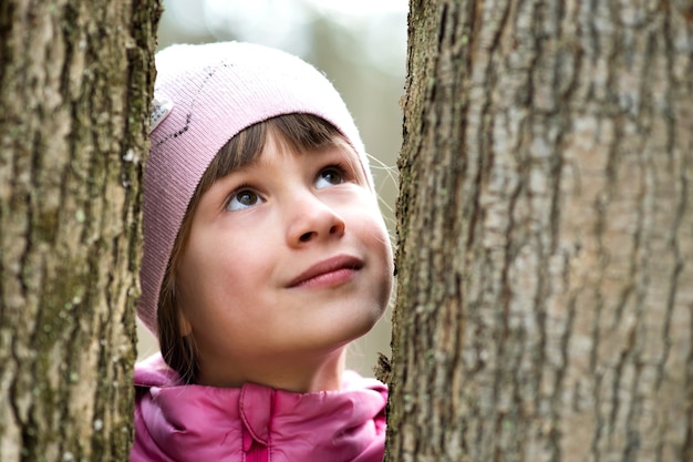 Portrait of young pretty child girl wearing pink jacket and cap standing between trees in park