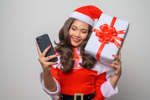 Портрет молодой красивой женщины в красном костюме санта-клауса, счастливого использовать смартфон с рождественской подарочной коробкой