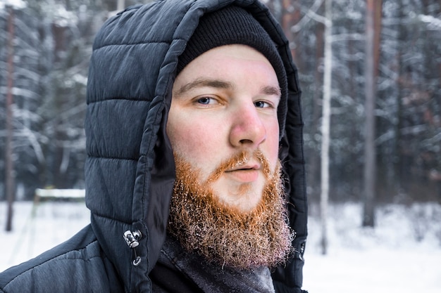 Portrait of a young red bearded European man in winter jacket and hat walks in a snowy winter park.