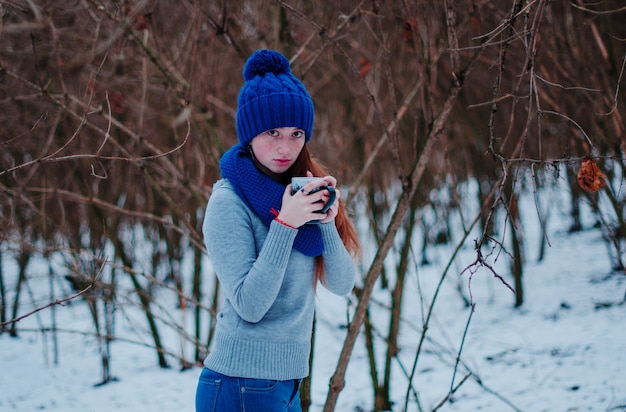 Portrait of young red hair girl with freckles wearing at blue knitted wool hat and scarf with cup of tea in winter day.