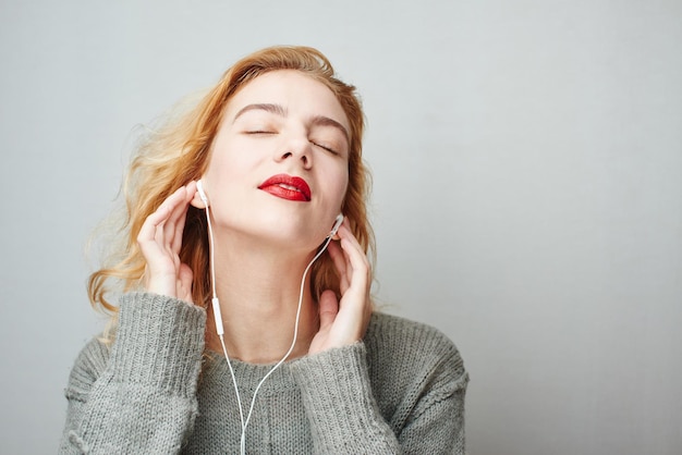 Portrait of a young red-haired girl on a gray background in a sweater listening to music with headphones