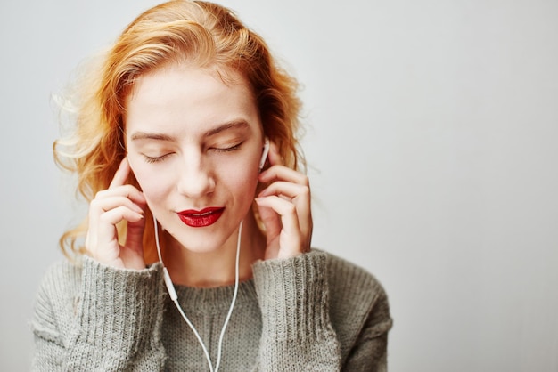 ヘッドフォンで音楽を聴いているセーターの灰色の背景に若い赤毛の少女の肖像画