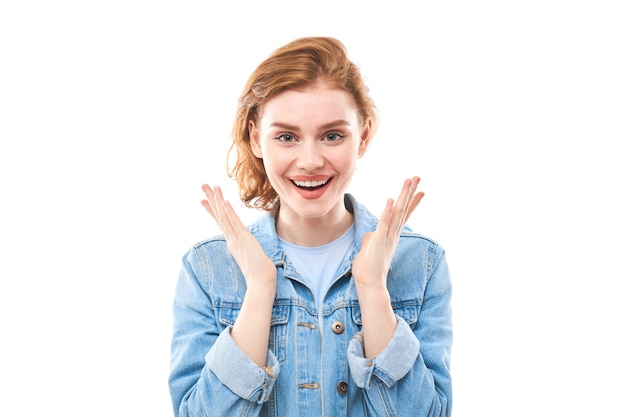 Portrait of a young red-haired girl on a white isolated background in jeans. Looks into the camera holding hands in front of you in a fist