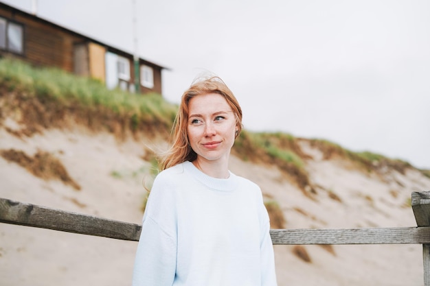 Portrait of young red haired woman in light blue sweater on sand beach by sea in storm