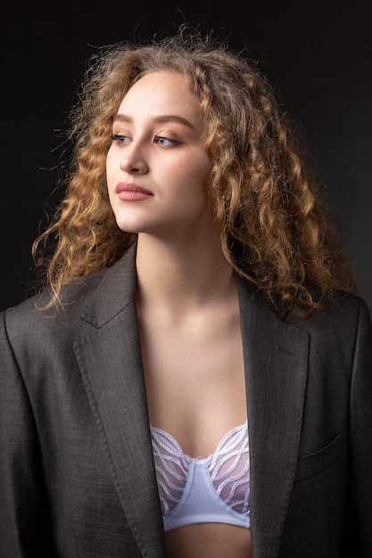 Portrait of a young redhaired curly sexy girl in a jacket with long hair in the studio Shows off her curvaceous breasts in a bra