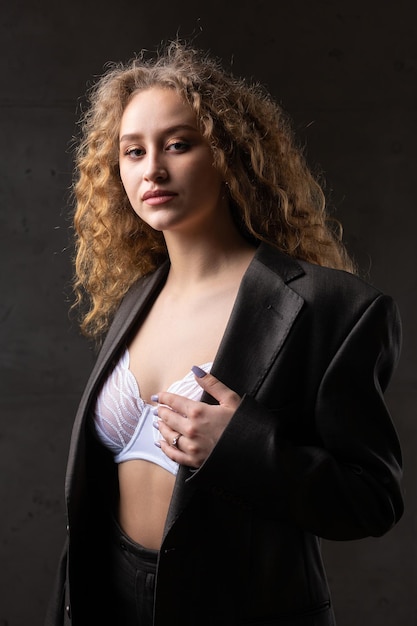 Portrait of a young redhaired curly sexy girl in a jacket with long hair in the studio Shows off her curvaceous breasts in a bra