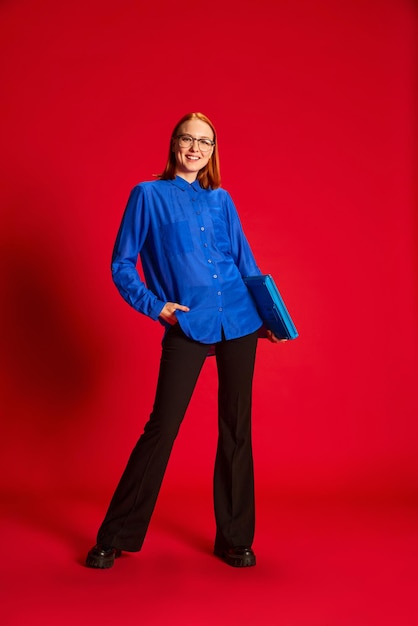 Portrait of young redhead girl in blue shirt and glasses posing with laptop over red background Motivated worker