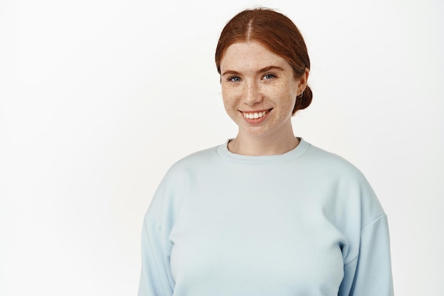 Portrait of young redhead girl with pale skin, white beautiful smile and freckles on face, looking happy and confident at camera, standing in casual outfit against white background