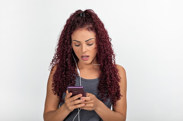 Portrait of a young redhead using cellphone with headphones, surprised and looking at the screen. Isolated