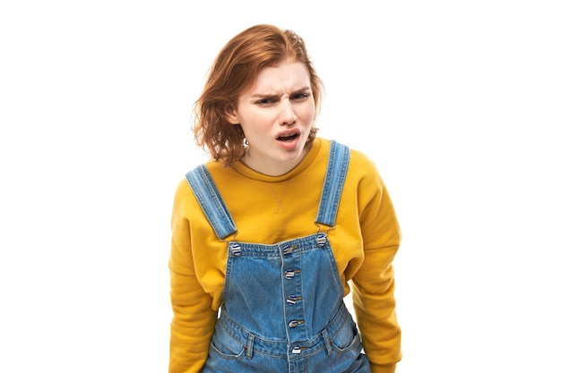 Portrait of young redhead woman in yellow casual suspiciously looking at camera isolated on white studio background squinting incredulously