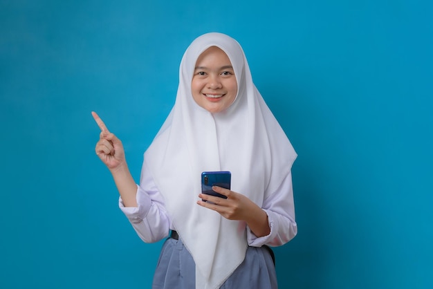 Портрет молодой расслабленной улыбающейся студентки в хиджабе, держащей мобильный телефон на синем фоне