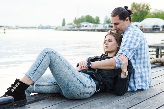 Portrait of young sensual and loving couple sitting on a pier beside a river