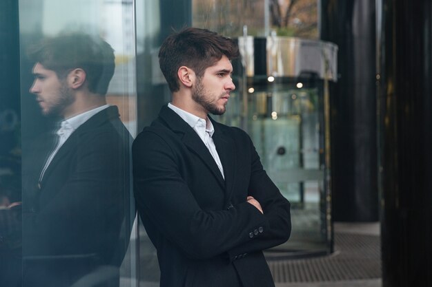 portrait of young serious businessman dressed in official clothing standing near business center and look aside.