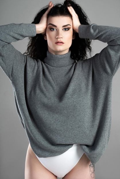 Portrait of young and sexy woman with black wavy hair wearing turtleneck jumper posing on gray background in studio