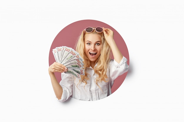 Portrait of a young shocked woman with a bundle of banknotes and text Sale. Peeping through the white hole  Funny face with open mouth. Wow concept