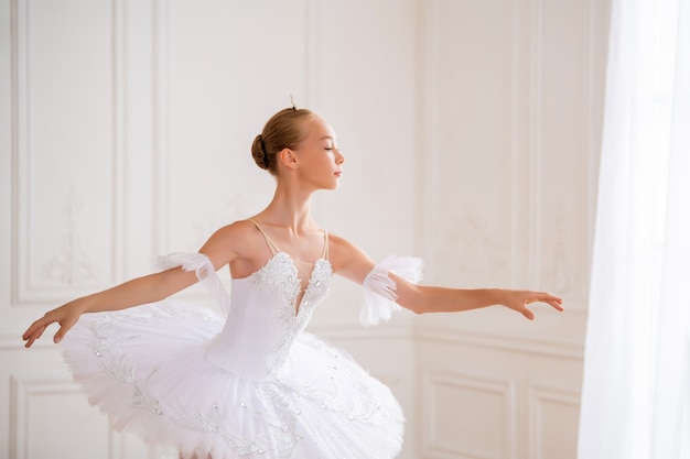 Portrait of a young slender ballerina in a white tutu in an elegant pose in a large beautiful white hall.