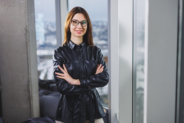 Portrait of a young slender woman in glasses and a black leather shirt Modern woman on the background of the window in the office with a large window