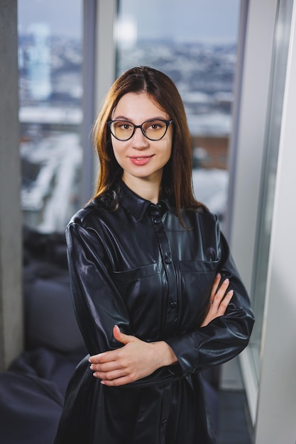Photo portrait of a young slender woman in glasses and a black leather shirt modern woman on the background of the window in the office with a large window