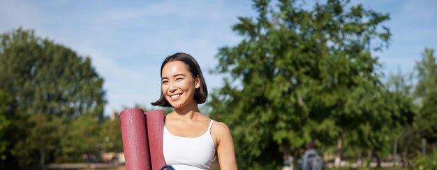 Photo portrait of young slim and healthy korean girl doing workout in park standing with water bottle and