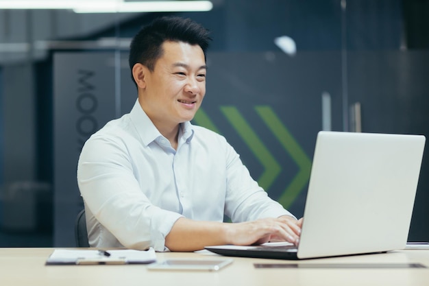 Портрет молодого улыбающегося азиатского бизнесмена, работающего в офисе, сидящего за столом и печатающего на
