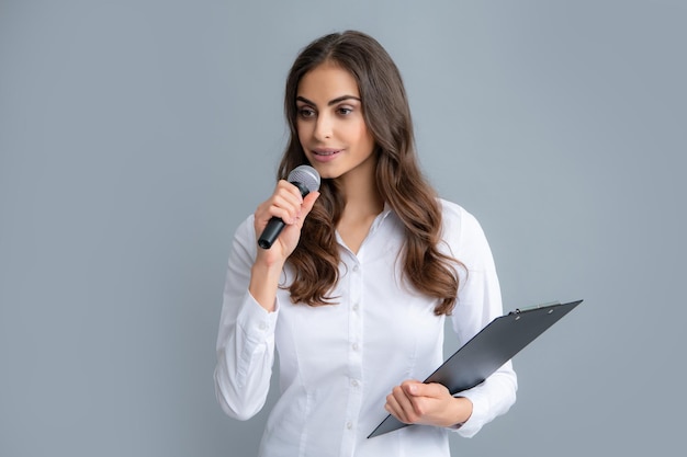 Portrait of young smiling business woman doing presentation with clipboard and microphone isolated grey background