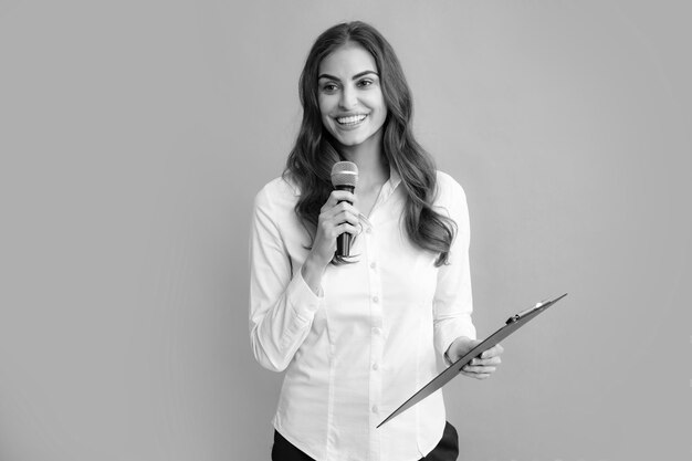 Portrait of young smiling business woman doing presentation with clipboard and microphone isolated grey background