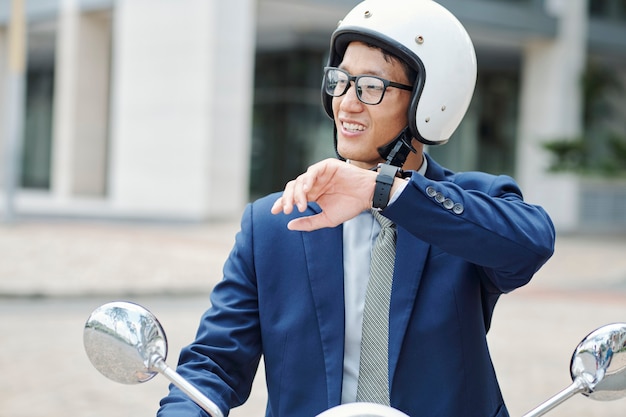 Portrait of young smiling businessman in helmet riding on scooter and recording voice message via application on smartwatch
