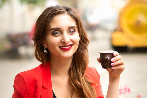 Портрет молодой улыбающейся кавказской женщины, смотрящей в камеру, сидя с чашкой кофе на открытом воздухе