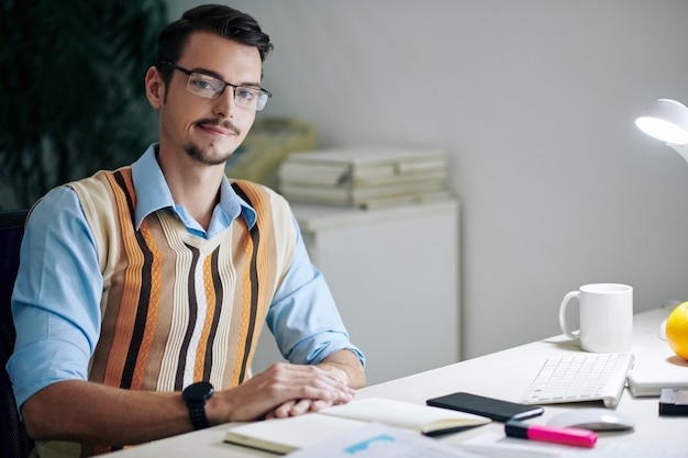 Portrait of young smiling entrepreneur in glasses sitting at office desk with opened planner and cup of coffee and smiling at camera