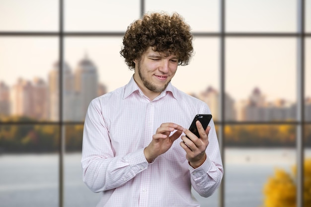 Портрет молодого улыбающегося мужчины с кудрявыми волосами нажимает на окно своего смартфона с городской рекой