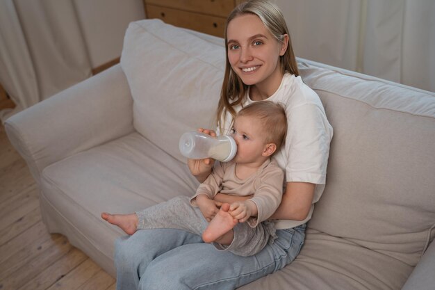 Portrait of young smiling mother feeding cute baby from bottle at home