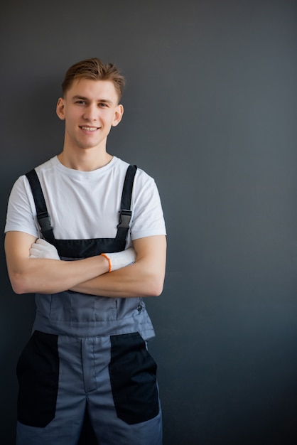 Portrait of a young, smiling, professional worker in gray overalls and white T-shirt, standing with arms crossed on a gray background.