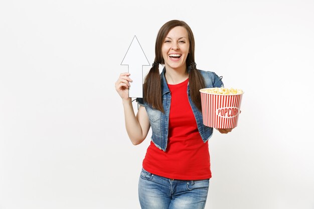 Portrait of young smiling woman in casual clothes watching movie film, holding bucket of popcorn, pointing white arrow up on copyspace isolated on white background. Emotions in cinema concept.