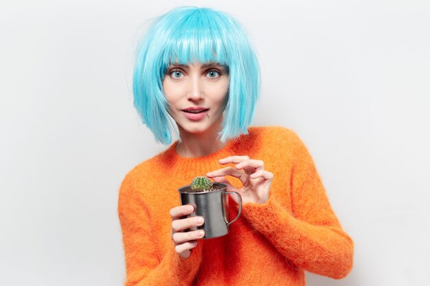 portrait of young smiling woman holding steel mug with cactus. Wearing blue wig and orange sweater.