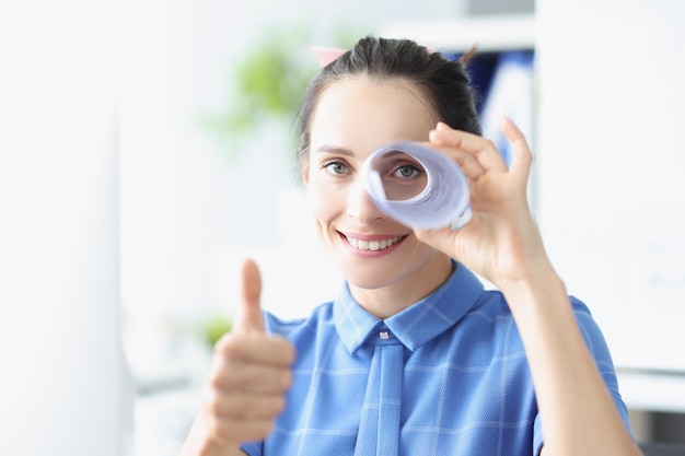 Portrait of a young smiling woman holds thumbs up and looks into pipe made of paper search for