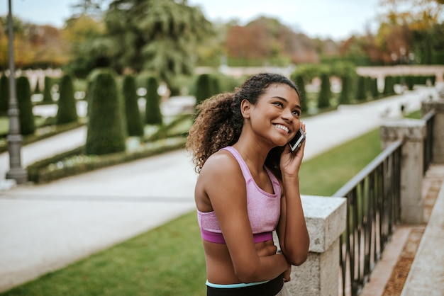 Ritratto degli abiti sportivi d'uso sorridenti della giovane donna che parlano sul telefono all'aperto.