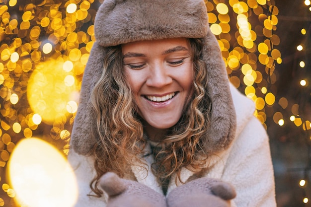 Portrait of young smiling woman with curly hair in fur hat in the winter street decorated with lights