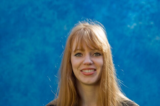 Portrait of young smiling woman with long blond hair against blue wall