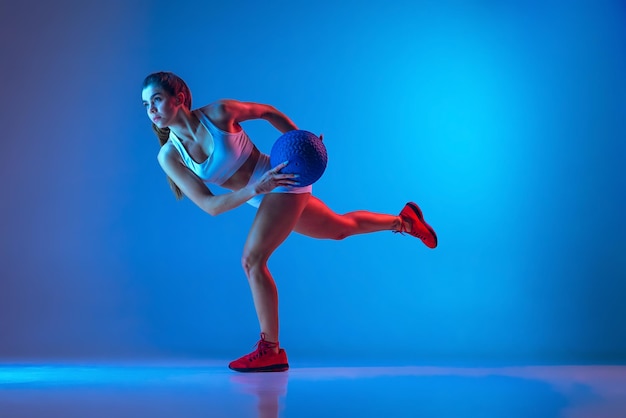 Ritratto di giovane ragazza sportiva che si allena in palestra facendo esercizi con palla isolata su sfondo blu al neon