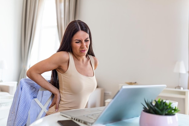 portrait of young stressed woman sitting at home office desk in front of laptop touching aching back with pained expression suffering from backache after working on laptop