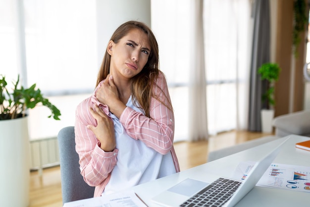 Photo portrait of young stressed woman sitting at home office desk in front of laptop touching aching shoulder with pained expression suffering from shoulder ache after working on laptop