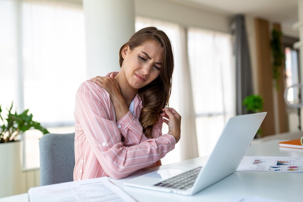 Portrait of young stressed woman sitting at home office desk in front of laptop touching aching shoulder with pained expression suffering from shoulder ache after working on laptop