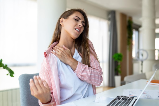 Portrait of young stressed woman sitting at home office desk in front of laptop touching aching shoulder with pained expression suffering from shoulder ache after working on laptop