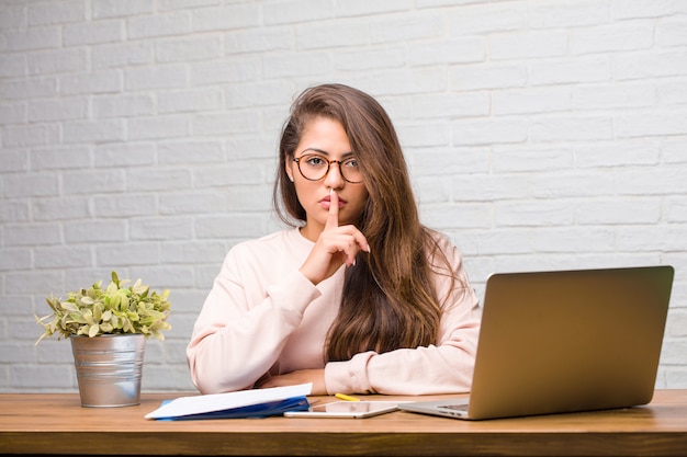 Portrait of young student latin woman sitting on her desk keeping a secret or asking for silence