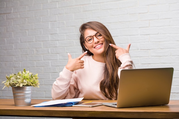 Portrait of young student latin woman sitting on her desk smiles, pointing mouth