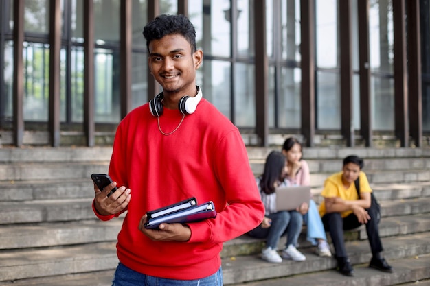 Портрет молодого студента, счастливо улыбающегося, держащего смартфон и книги в университете