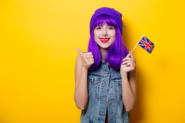 Portrait of young style hipster girl with purple hair and Great Britain flag in hand on yellow background