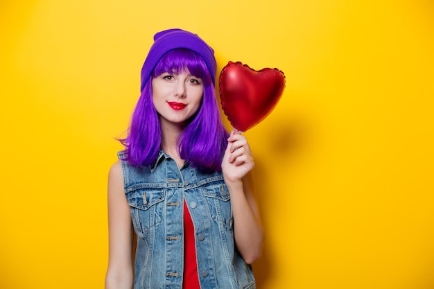 Portrait of young style hipster girl with purple hair and heart shape balloon on yellow background. st. valentines day holiday