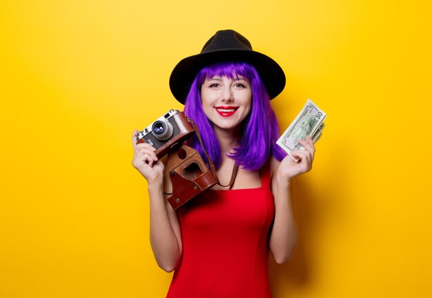 Portrait of young style hipster girl with purple hairstyle with money and retro camera on yellow background