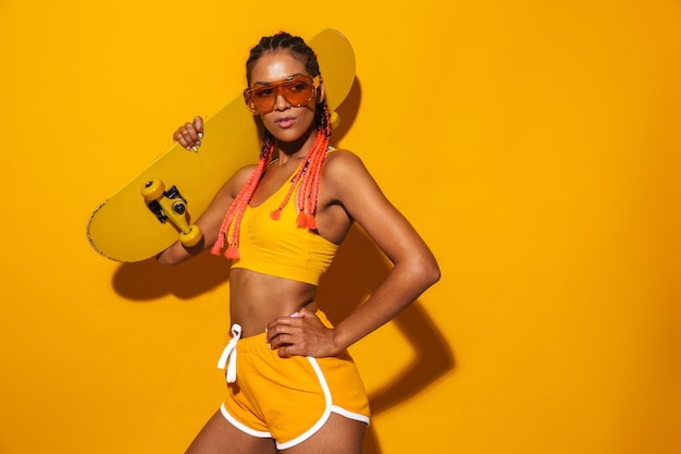 Portrait of a young stylish african woman wearing sports clothing standing isolated over yellow wall, holding skateboard